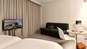 Una habitación de hotel moderna con un televisor de pantalla plana grande en la pared, un sofá negro, un sillón blanco, una mesita redonda y una maleta marrón en el suelo.