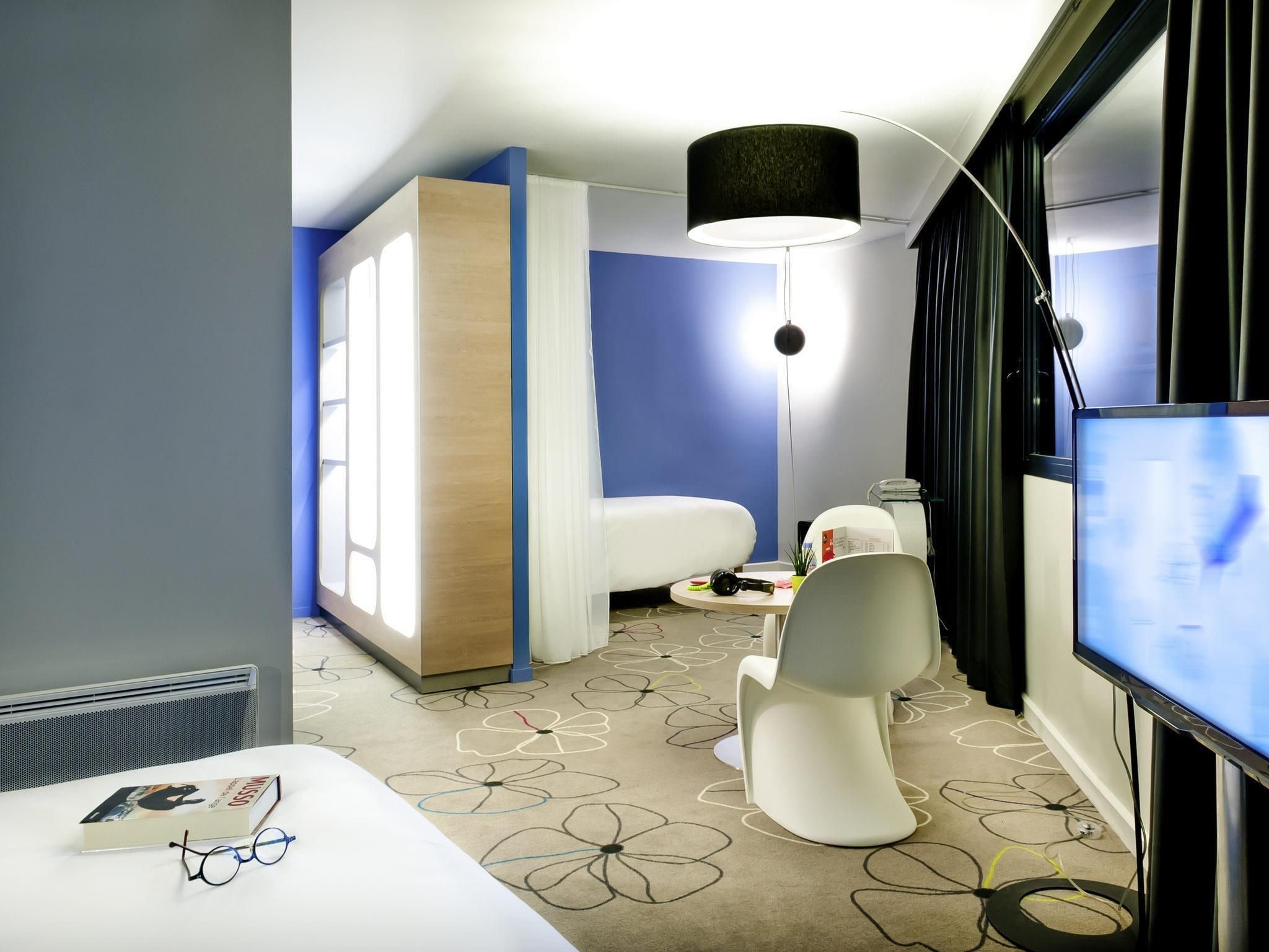 A modern hotel room with a bed, a desk with a computer, a chair, a TV, and a large window.
