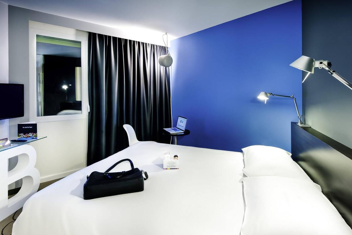 A hotel room with a bed, a desk with a laptop, a lamp, and a window with curtains.