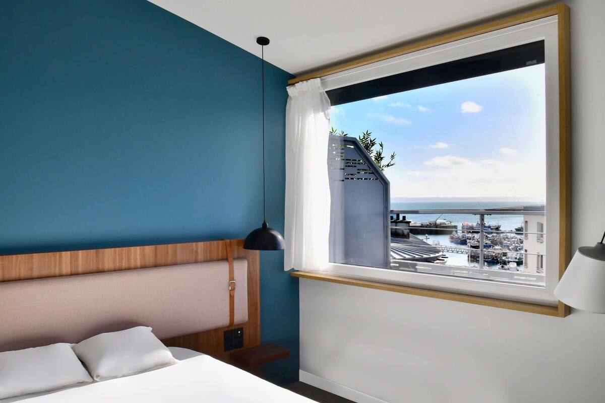 Habitación de hotel con cama, mesita de noche, lámpara y ventana con vistas al puerto deportivo.