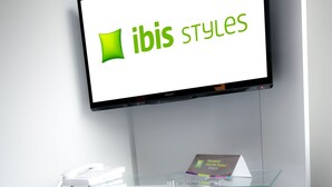 A modern reception desk with a glass top, a mounted television displaying 'ibis styles', and a white 'B' shaped base. - ibis Styles Brest Centre Port (Brest)