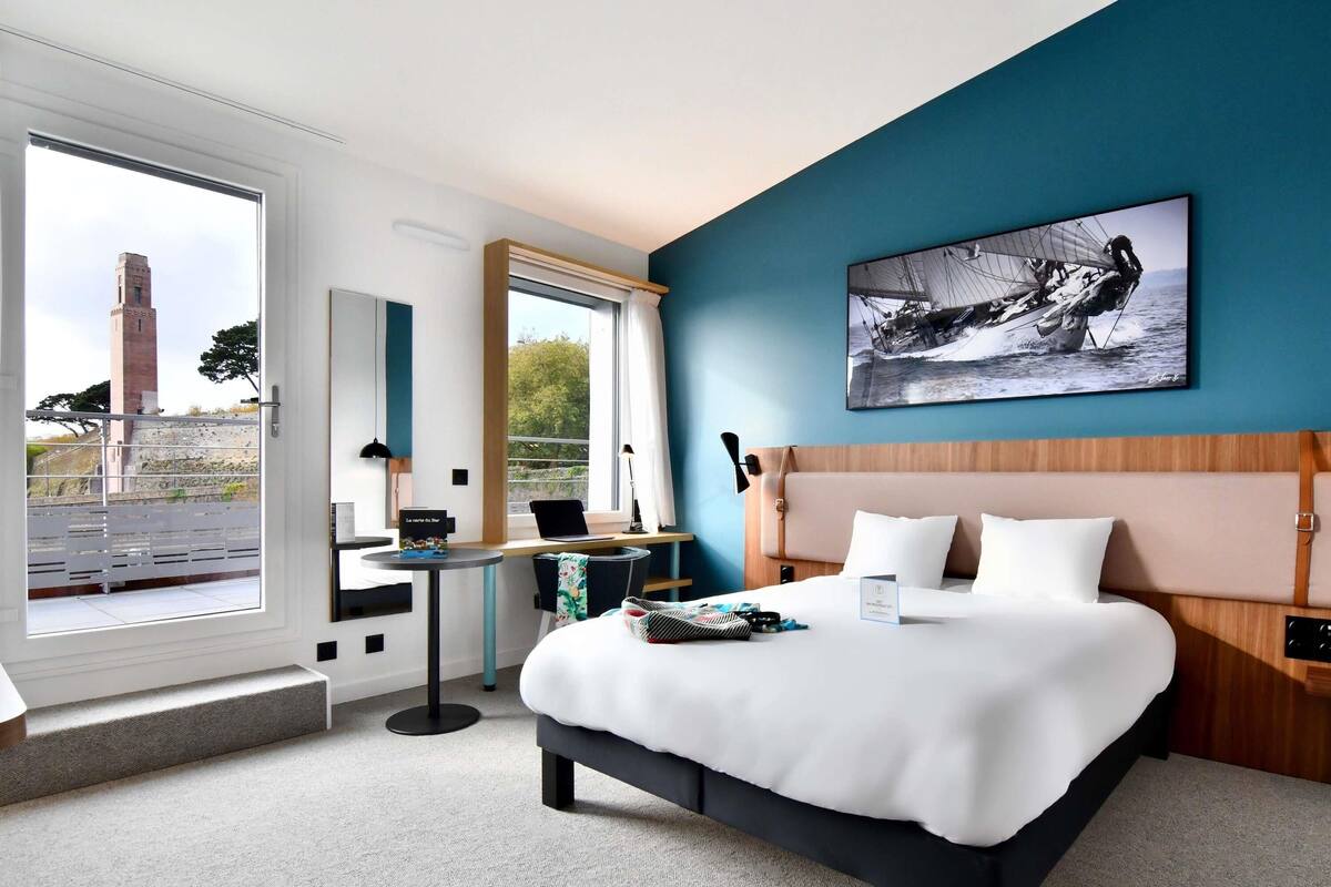 Una habitación de hotel con una cama, un escritorio, una computadora portátil, una ventana con vista a una torre y un cuadro en la pared.