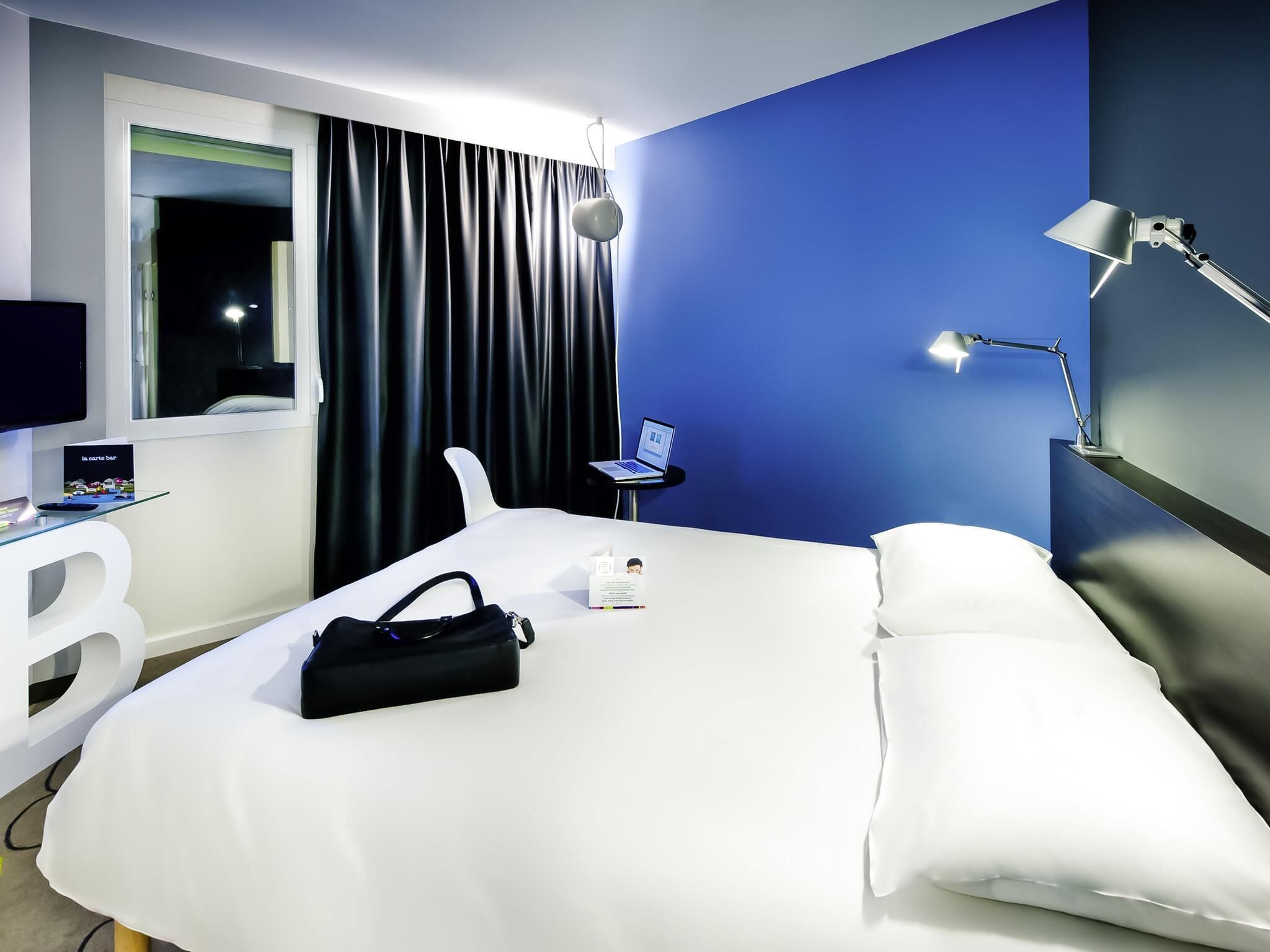 Une chambre d’hôtel avec un lit, un bureau avec un ordinateur portable, une lampe et une fenêtre avec des rideaux.