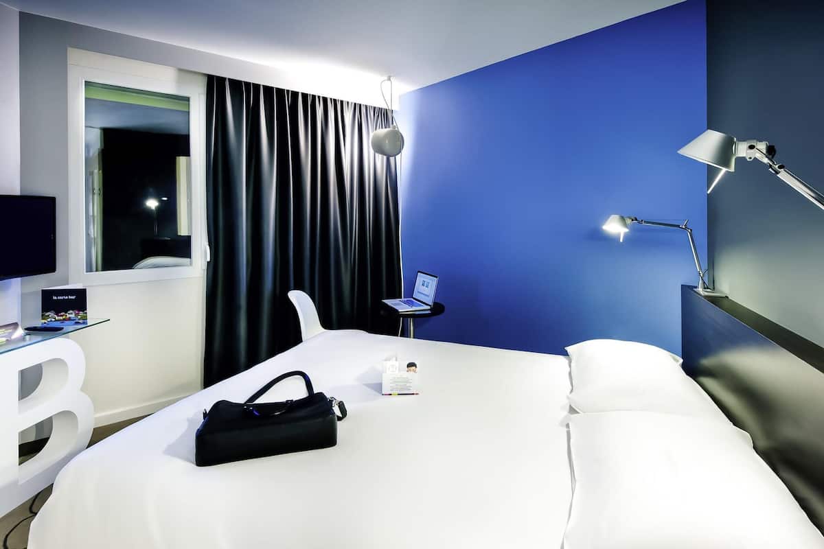 Une chambre d’hôtel avec un lit, un bureau avec un ordinateur portable, une lampe et une fenêtre avec des rideaux.