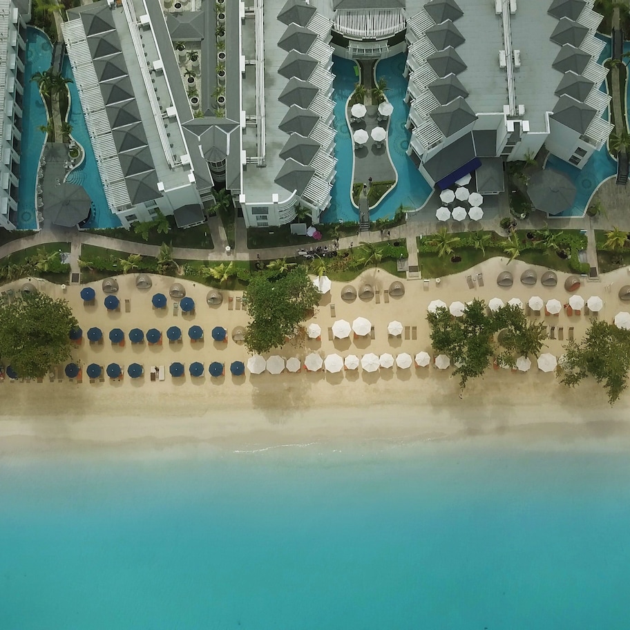 An aerial view of a resort with multiple buildings, a central swimming pool, and a beach area with umbrellas and lounge chairs.