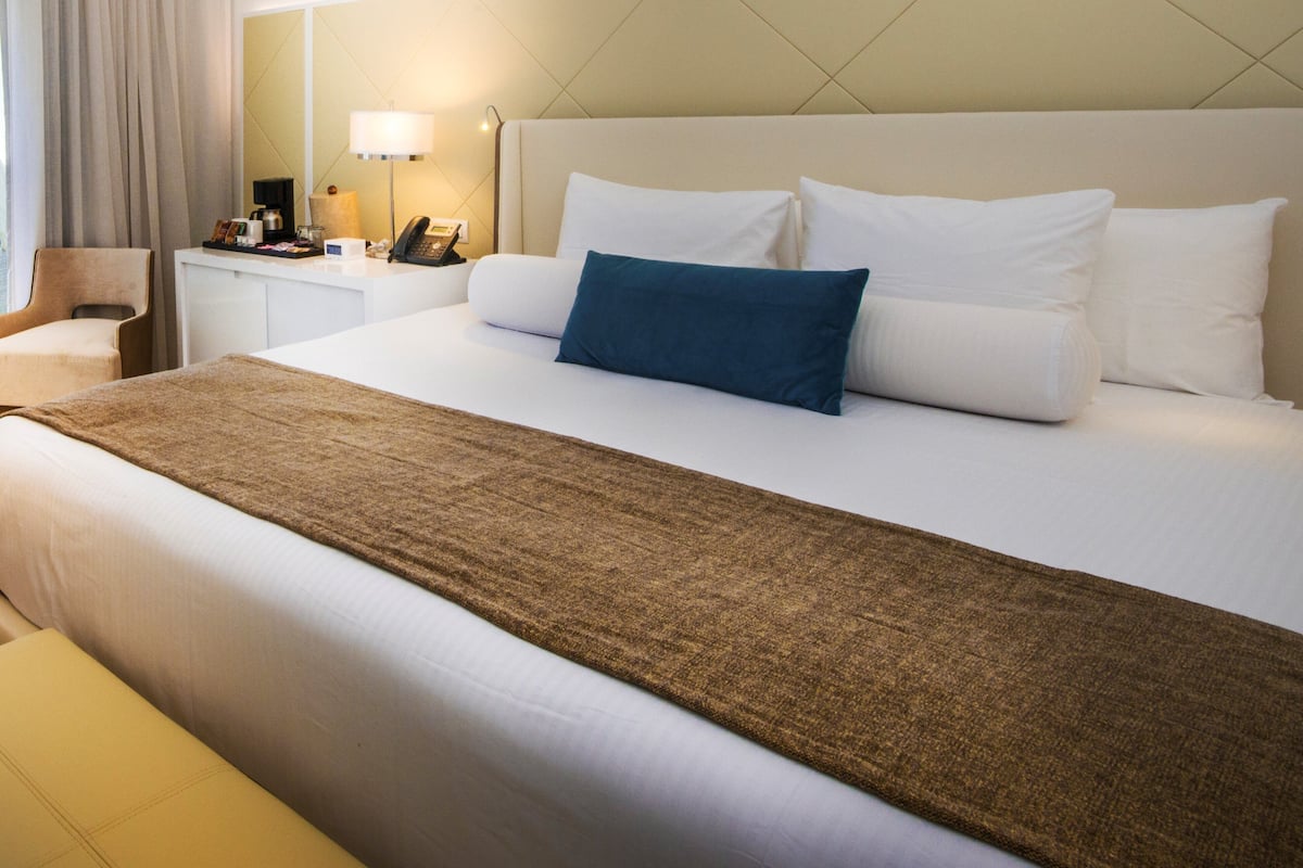 A neatly made bed with white and blue pillows, a brown throw blanket, and a bedside table with a lamp and phone.