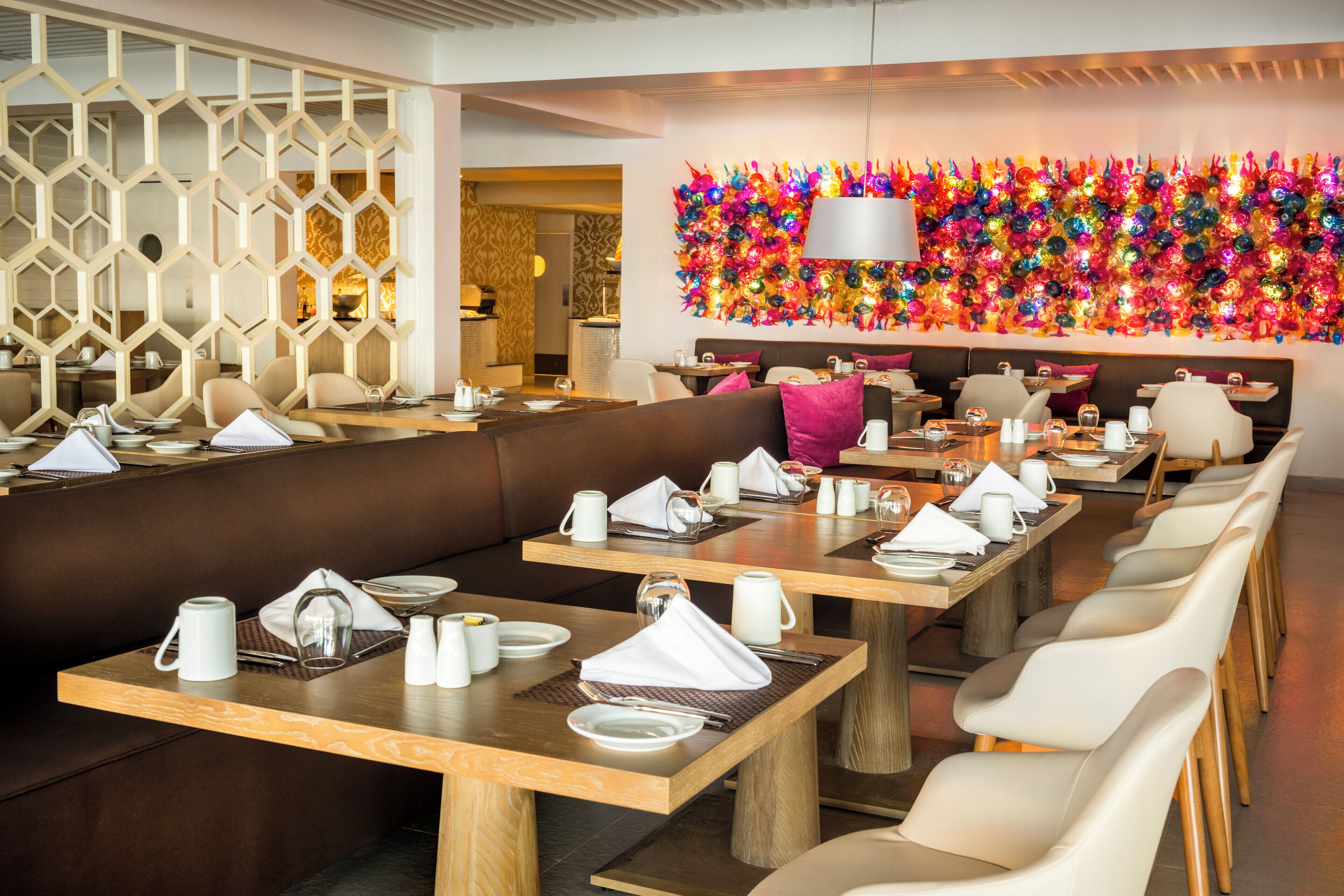 A modern restaurant with long communal tables, cushioned seating, and a vibrant decorative wall.