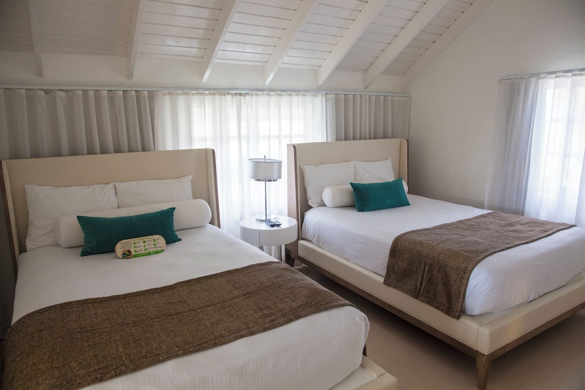 Two single beds with white linens and teal pillows, a small round table with a lamp, and a ceiling with exposed beams.