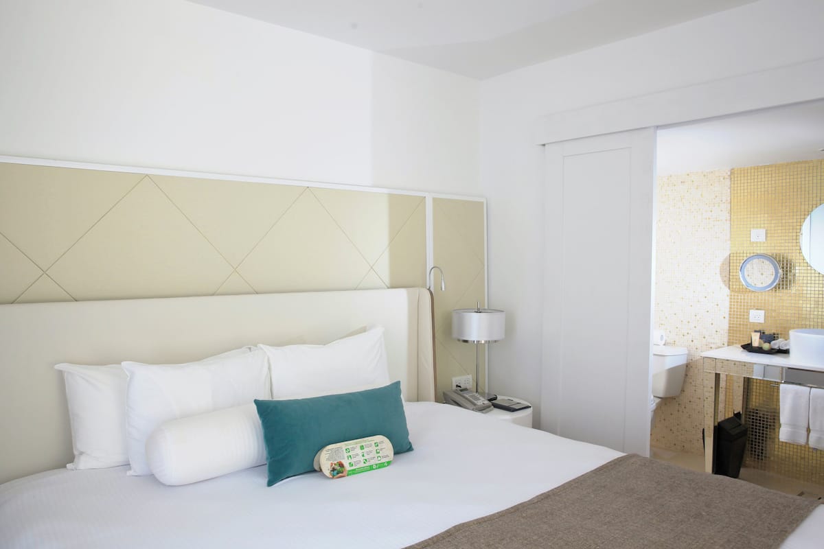 A hotel room with a bed, a bedside table with a lamp, a phone, and a bathroom with a sink and mirror.