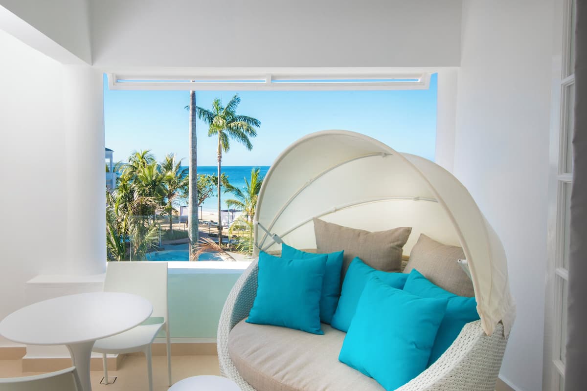 A balcony with a white wicker chair, a small round table, and a view of the beach and palm trees.