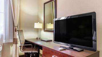 A hotel room with a large flat-screen TV, a wooden desk, a chair, a desk lamp, a mirror, and a window with curtains.
