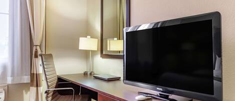 Hotelkamer met een groot flatscreen televisietoestel, een houten bureau, een stoel, bureaulamp, een spiegel en een raam met gordijnen.