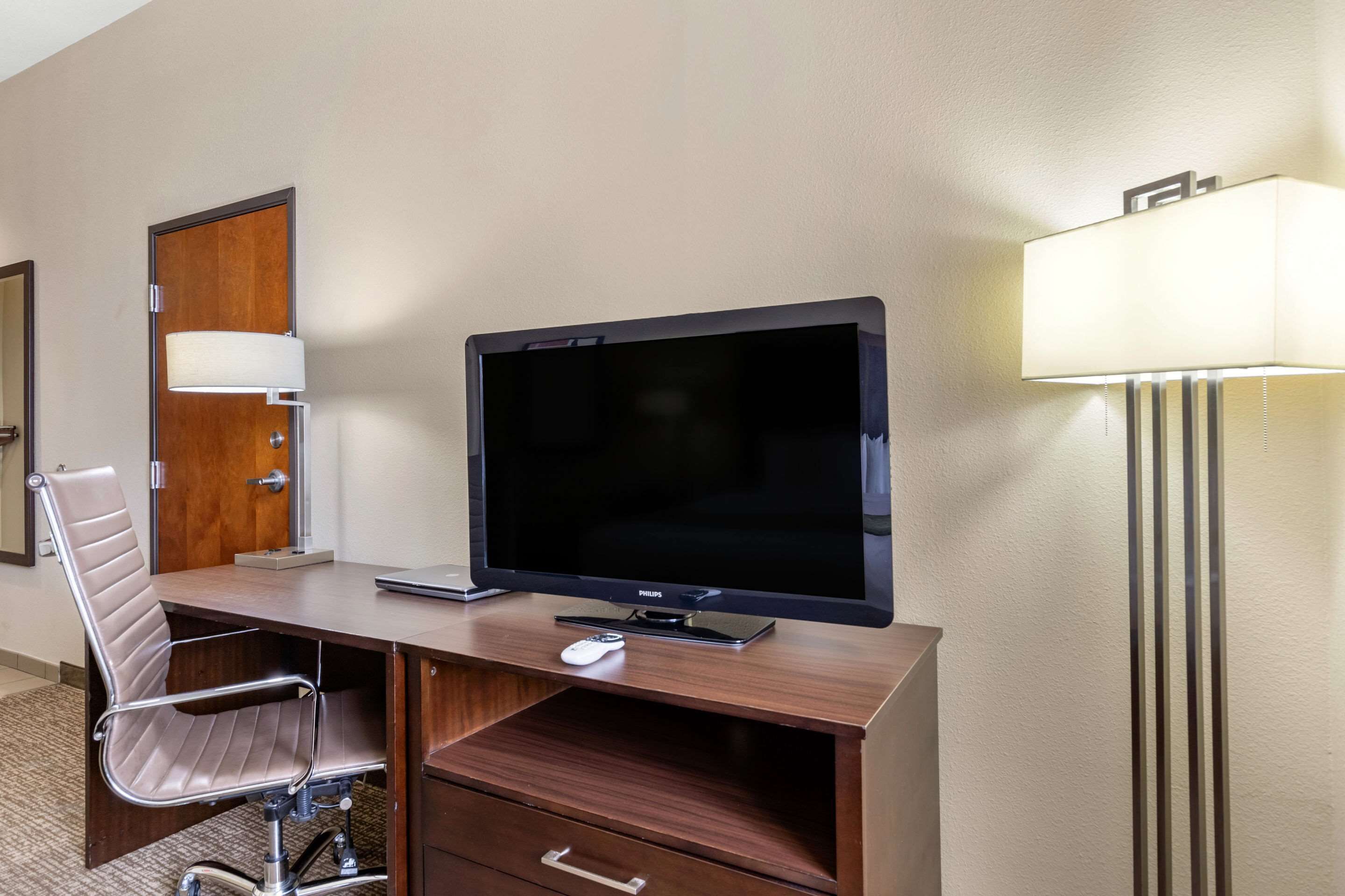 A hotel room with a wooden desk, a flat-screen TV, a modern chair, a desk lamp, and a door with a mirror.