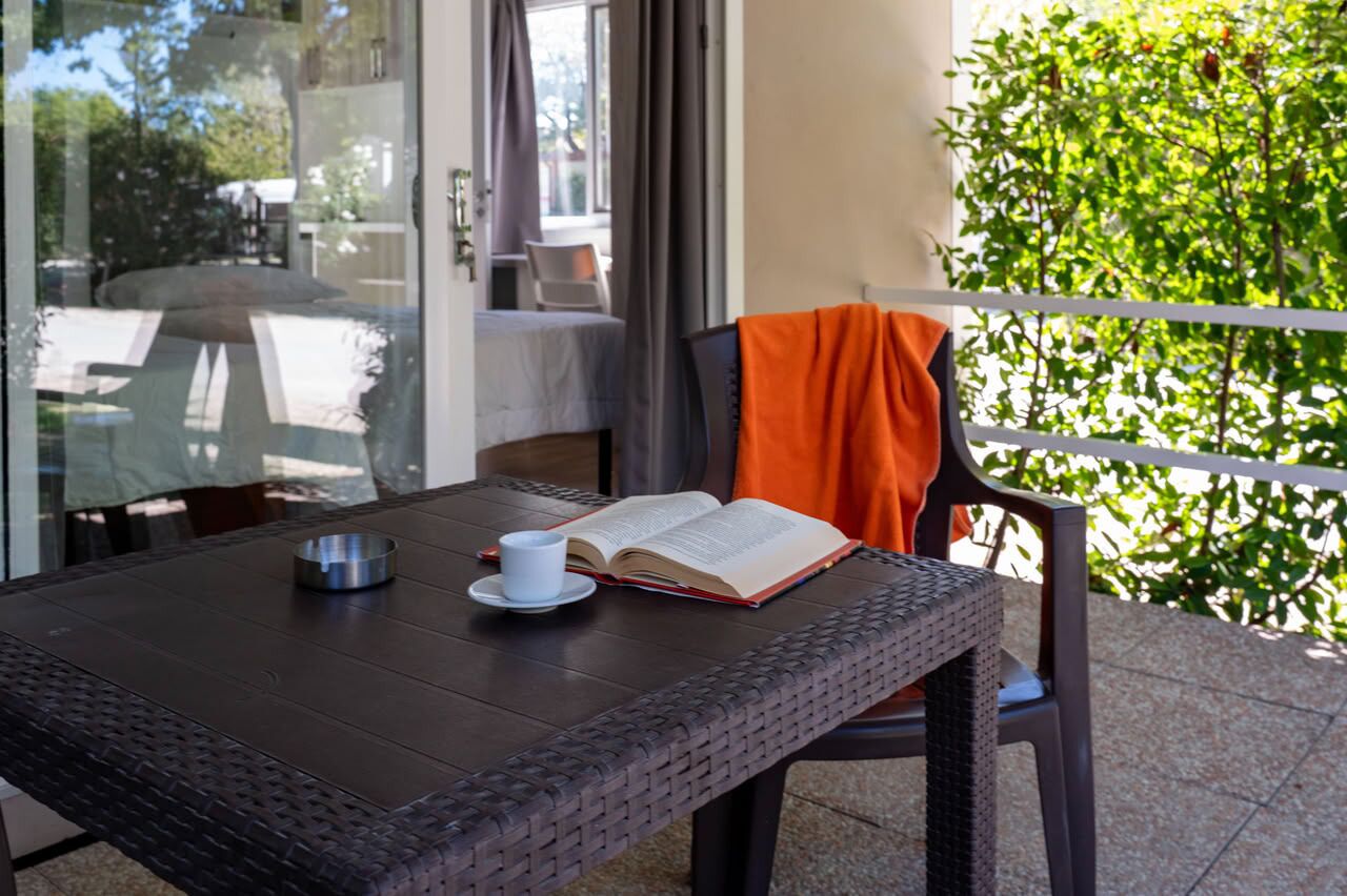Une table en osier sur laquelle se trouvent un livre et une tasse, une chaise avec une serviette orange, et une vue sur la verdure par une fenêtre.