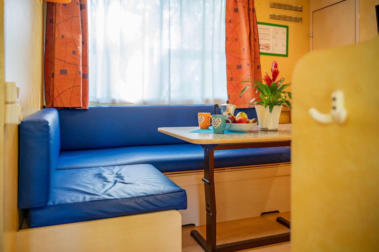Une pièce de taille modeste, avec un banc bleu rembourré, une petite table ornée d’une plante et d’un panier de fruits, et une fenêtre agrémentée de rideaux.