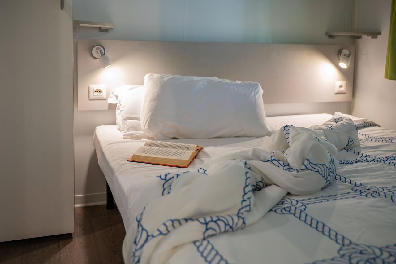 A neatly made bed with white bedding and blue patterns, a book on the bed, and a wall-mounted light fixture.