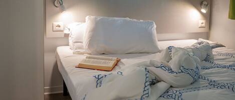 A neatly made bed with white bedding and blue patterns, a book on the bed, and a wall-mounted light fixture.