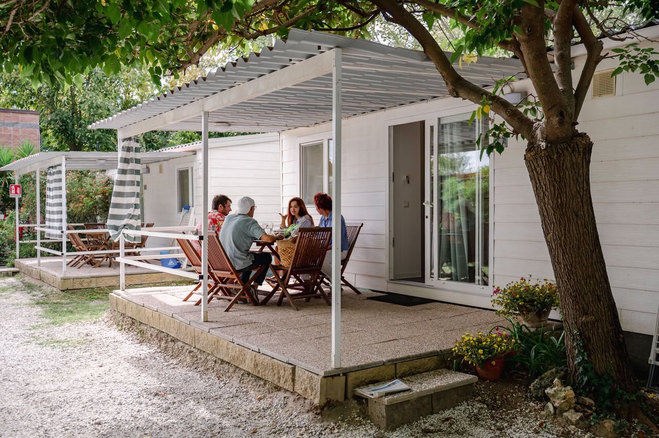 Ein überdachter Außenbereich mit Tisch und Stühlen, ein kleiner Garten mit Kübelpflanzen und ein Baum.