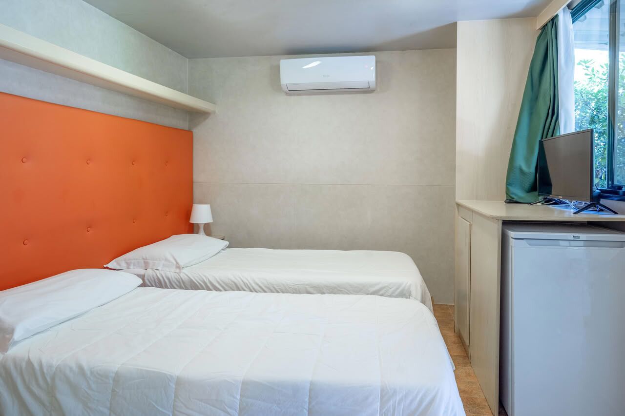 Una habitación de hotel con dos camas, un escritorio pequeño con una computadora, un refrigerador y un equipo de aire acondicionado en la pared.