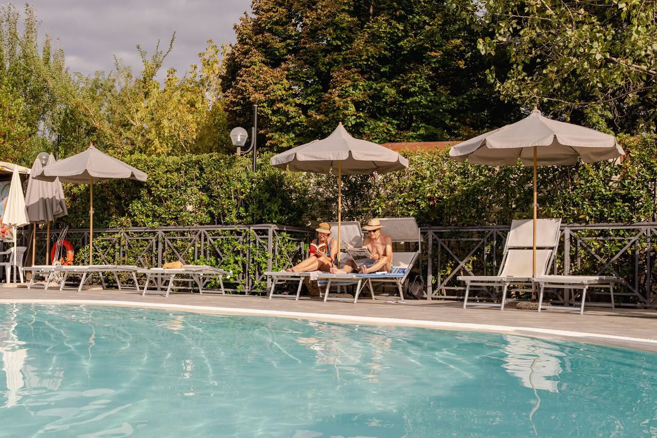 Zona de piscina con sillones de descanso y sombrillas, rodeada de follaje.