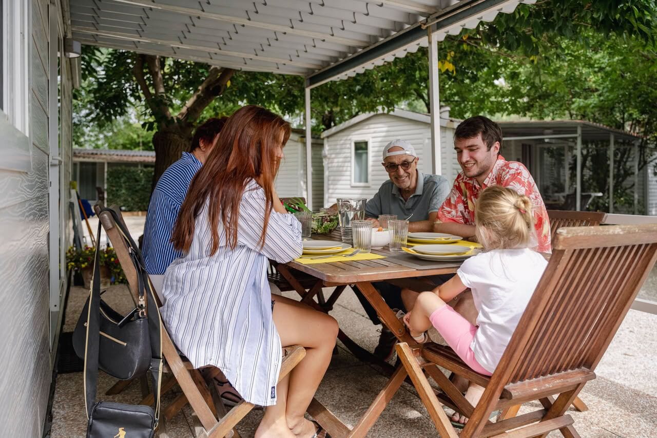 Una familia cenando en una mesa al aire libre, bajo un porche cubierto, rodeada de vegetación.