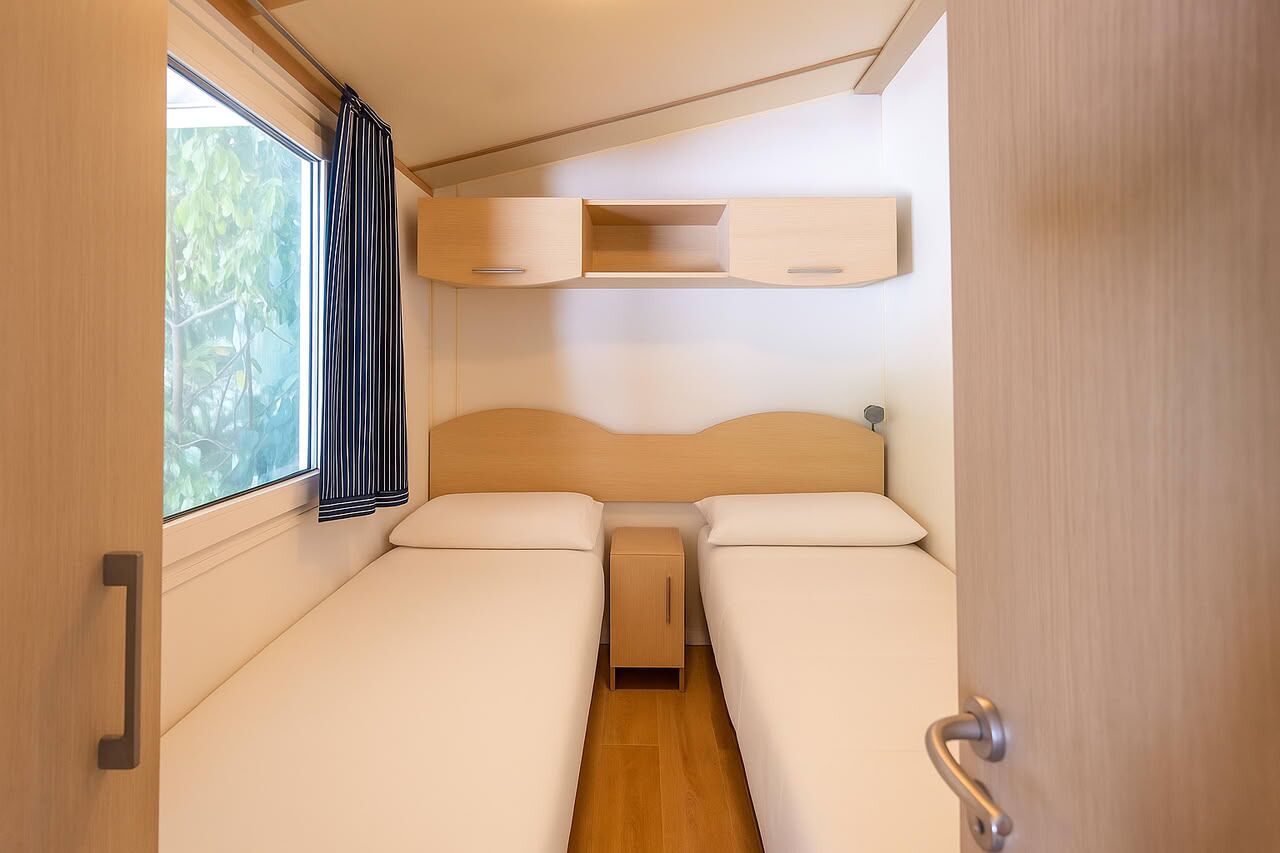 A compact RV bedroom with two beds, a small cabinet, and a window with curtains.