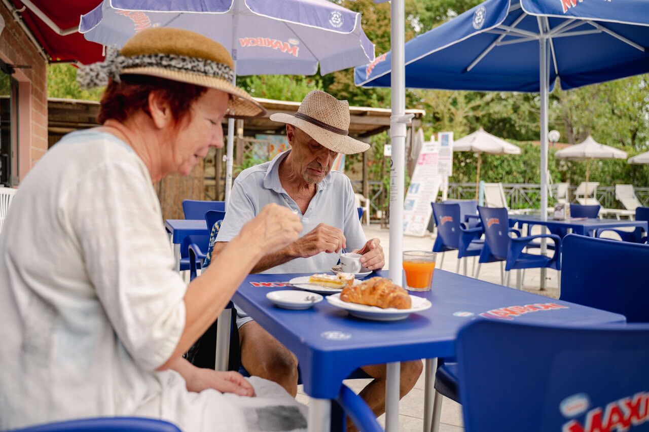 Two people dining at an outdoor table with umbrellas, chairs, and a sign in the background.