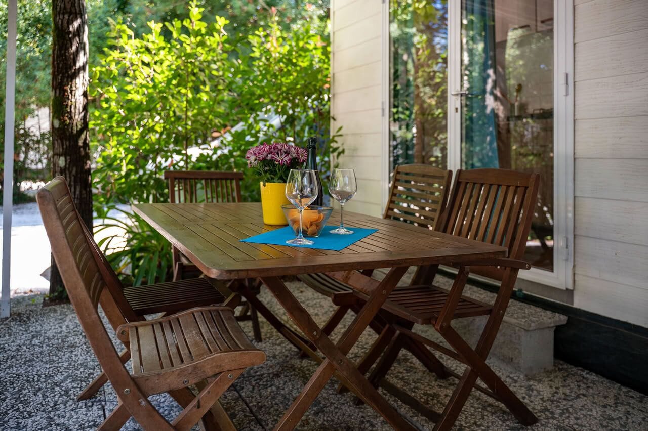 A wooden table with four chairs, a bottle, and two wine glasses on a blue mat, set outdoors with a garden view.