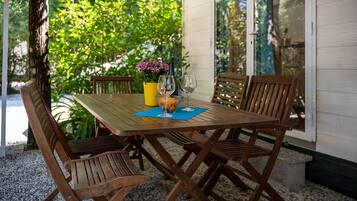 A wooden table with four chairs, a bottle, and two wine glasses on a blue mat, set outdoors with a garden view.