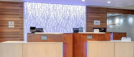Un hall d'accueil d'hôtel avec des panneaux de bois, deux comptoirs de réception, une grande fenêtre donnant sur une branche d'arbre et un écran numérique.