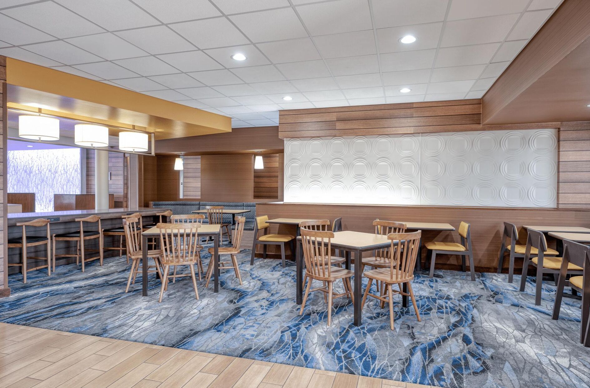 A hotel dining area with wooden tables and chairs, a patterned carpet, and a large decorative wall panel.