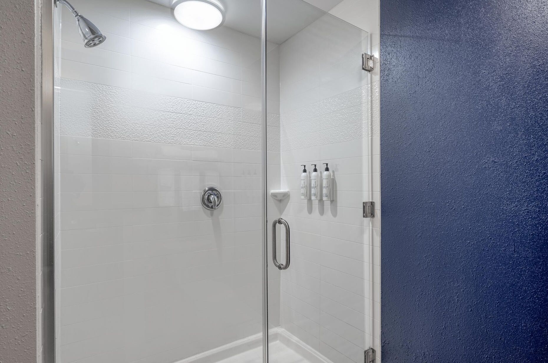 Um chuveiro envidraçado com azulejos brancos, um chuveiro manual e três frascos de shampoo na parede.