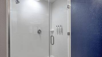 Um chuveiro envidraçado com azulejos brancos, um chuveiro manual e três frascos de shampoo na parede.