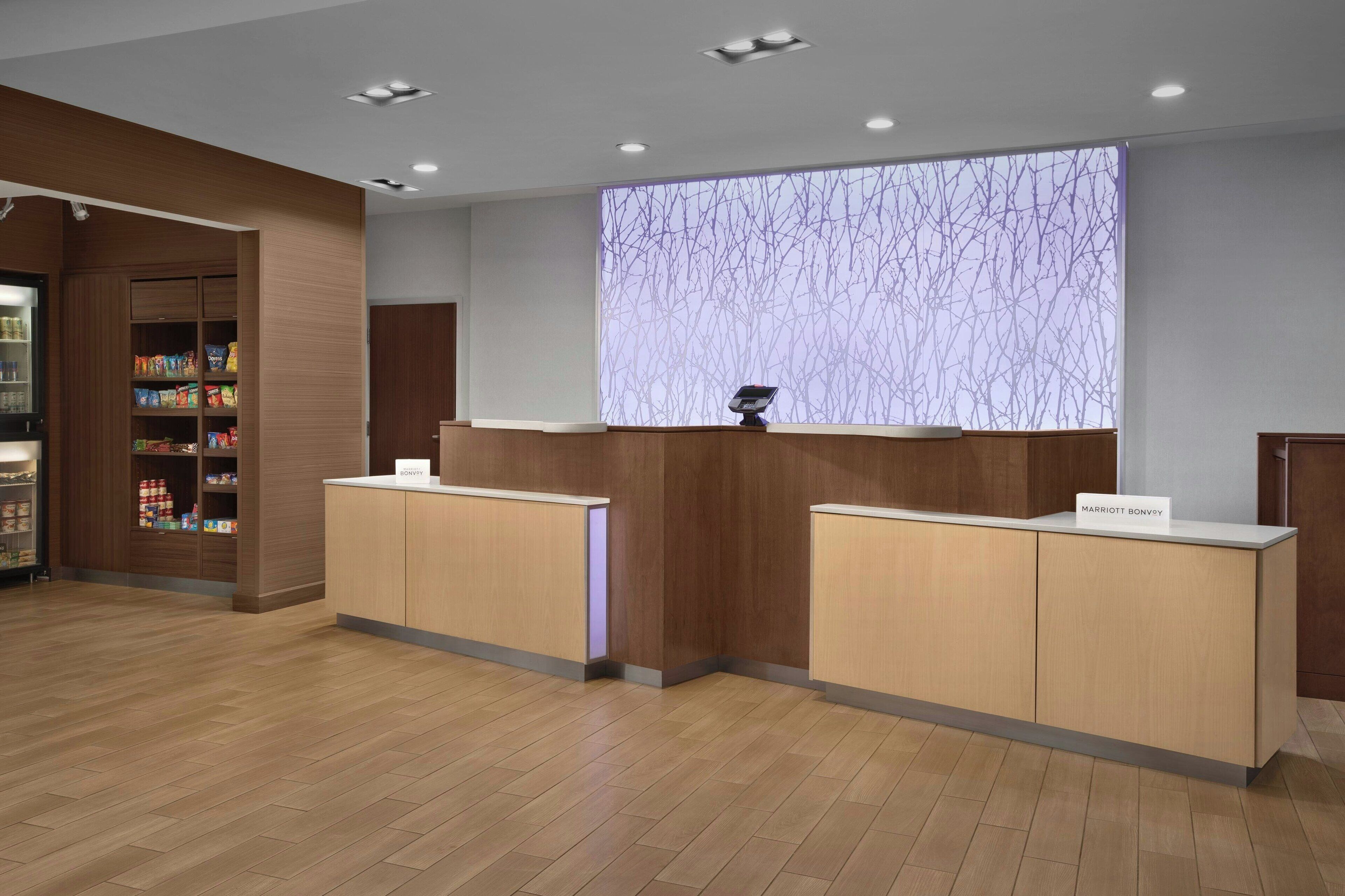 Un espace d’accueil d’hôtel avec des comptoirs de réception en bois, un écran numérique affichant un motif d’arbre et un bar servant des collations, où l’on propose divers aliments emballés.