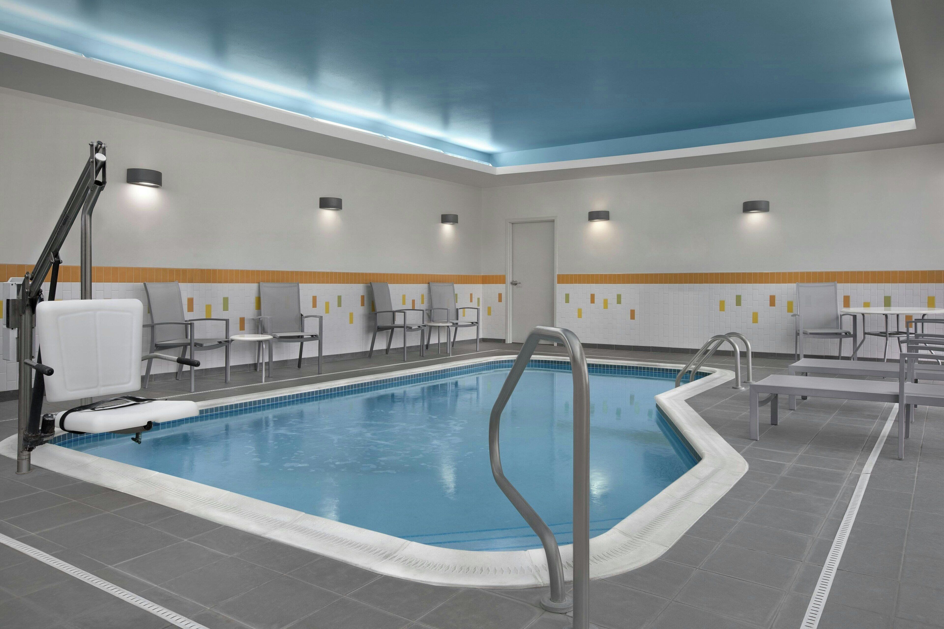 A swimming pool with a blue interior, surrounded by grey tiles, and equipped with a ladder and a bench. There are chairs and tables along the poolside, and the ceiling has recessed lighting.