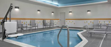 A swimming pool with a blue interior, surrounded by grey tiles, and equipped with a ladder and a bench. There are chairs and tables along the poolside, and the ceiling has recessed lighting.