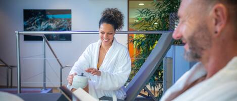A person in a white bathrobe sitting on a lounge chair in a modern indoor setting with a staircase and a plant in the background.