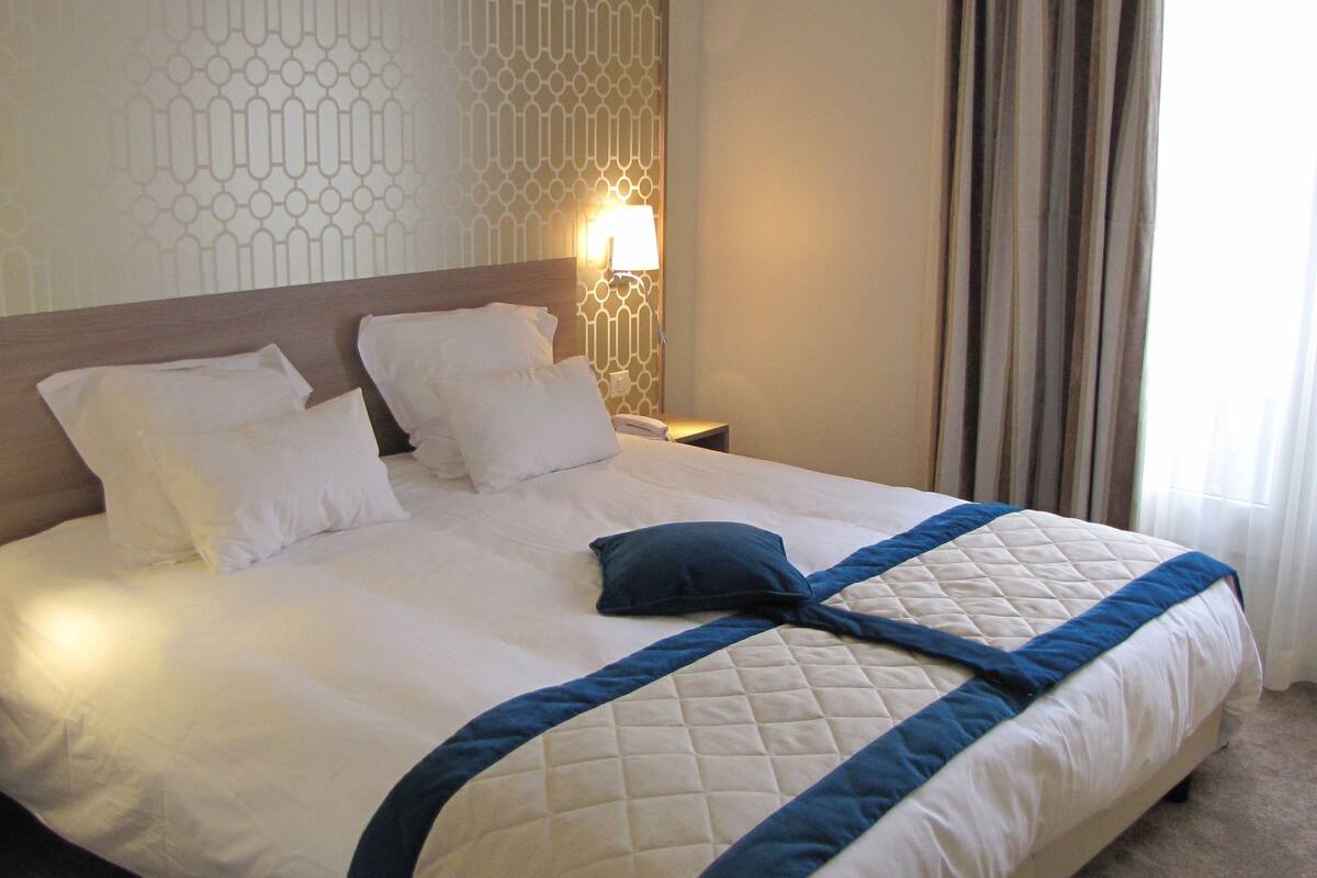 A neatly made bed with white linens and a blue accent, a bedside lamp, and a patterned wall.