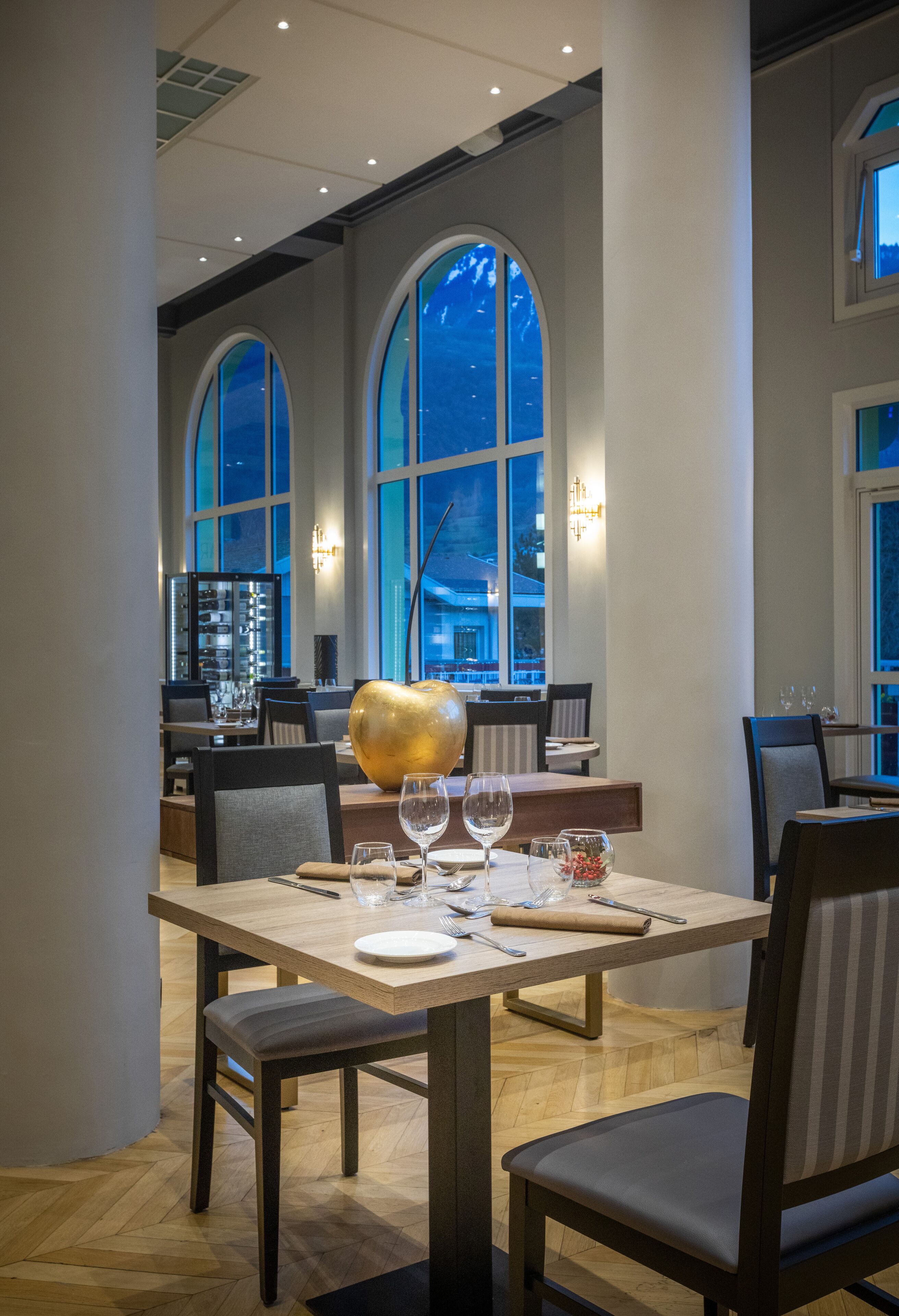 A dining area with a table set for two, large arched windows, and a view of the cityscape.