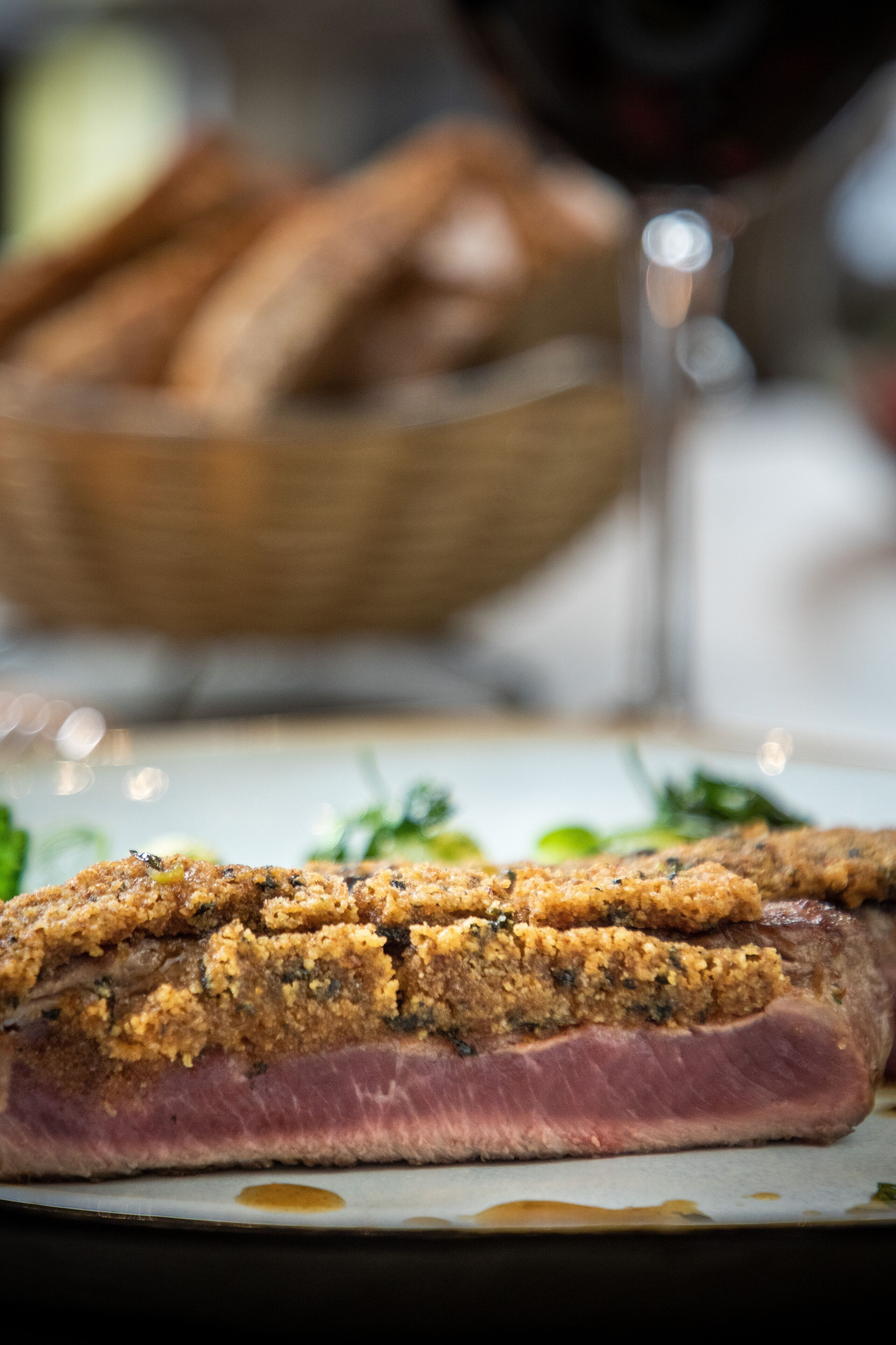 A close-up of a sliced meat dish with a crispy breadcrumb topping, a basket of bread, and a glass of red wine in the background.