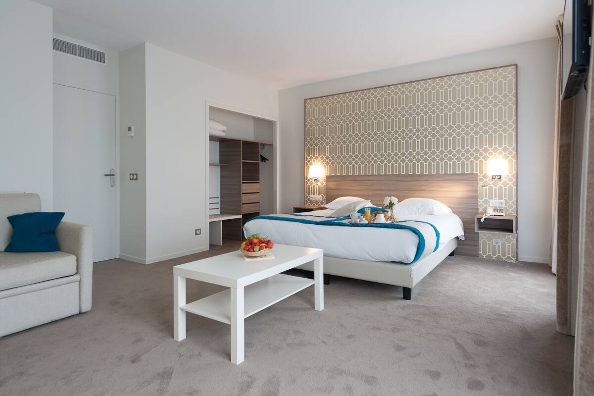 A modern hotel room with a bed, bedside tables, a small table with fruit, a sofa, and a wardrobe.