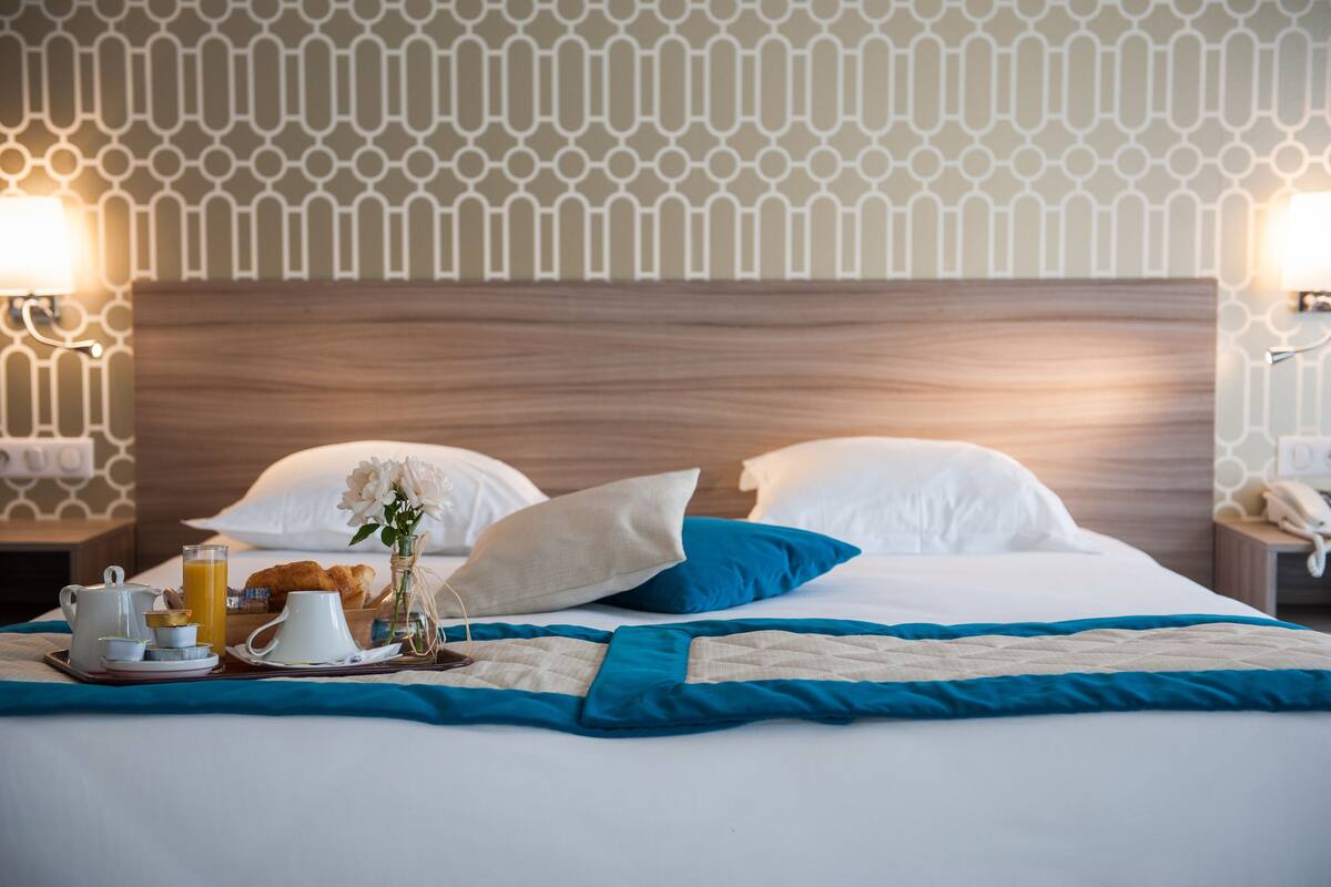 A neatly made bed with a blue and beige bedspread, white pillows, and a tray with breakfast items including croissants, coffee, and orange juice.