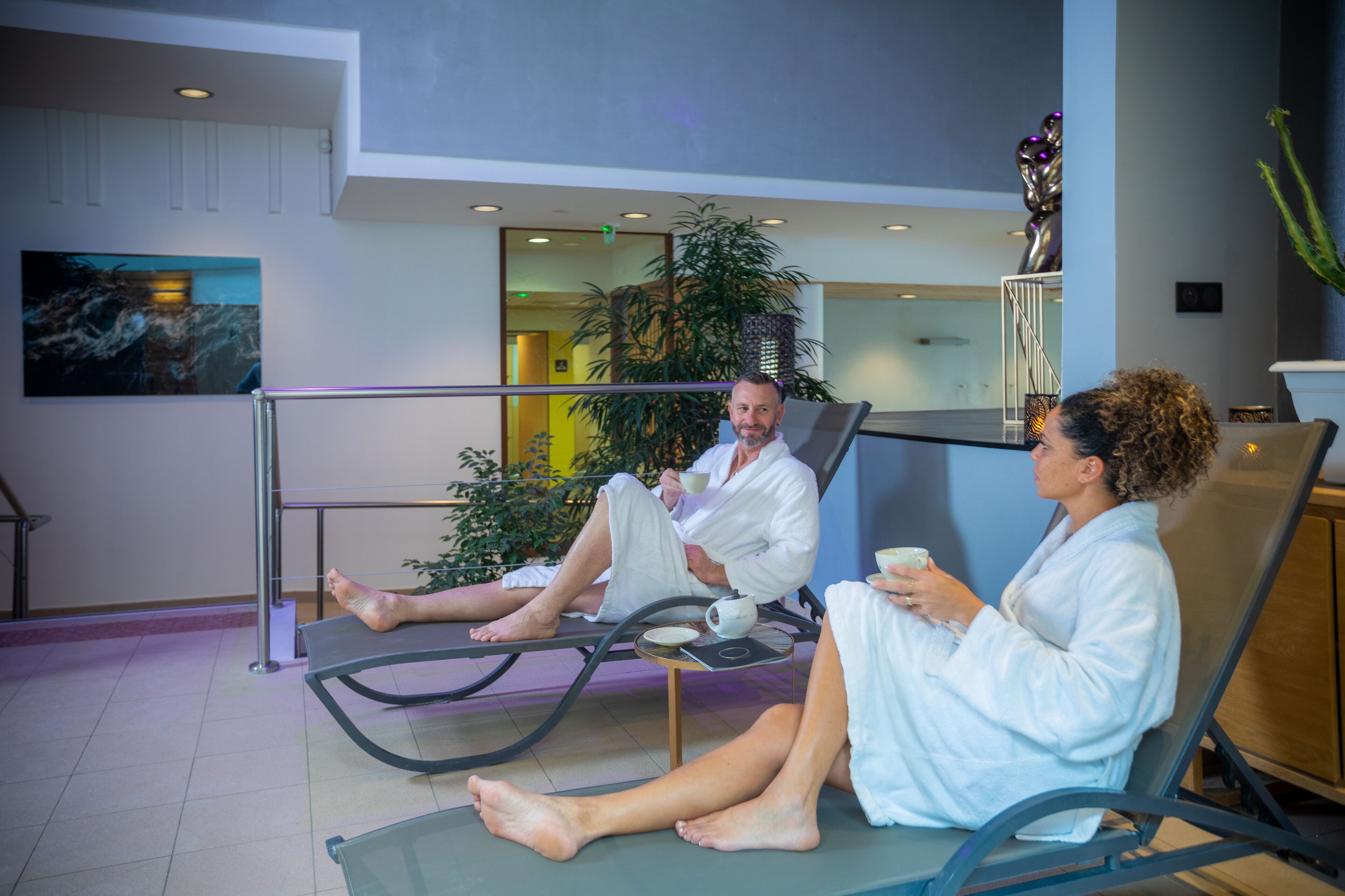 Dos personas vestidas de blanco sentadas en sillones reclinables en un ambiente interior moderno con barandilla de cristal, una planta y un cuadro en la pared.
