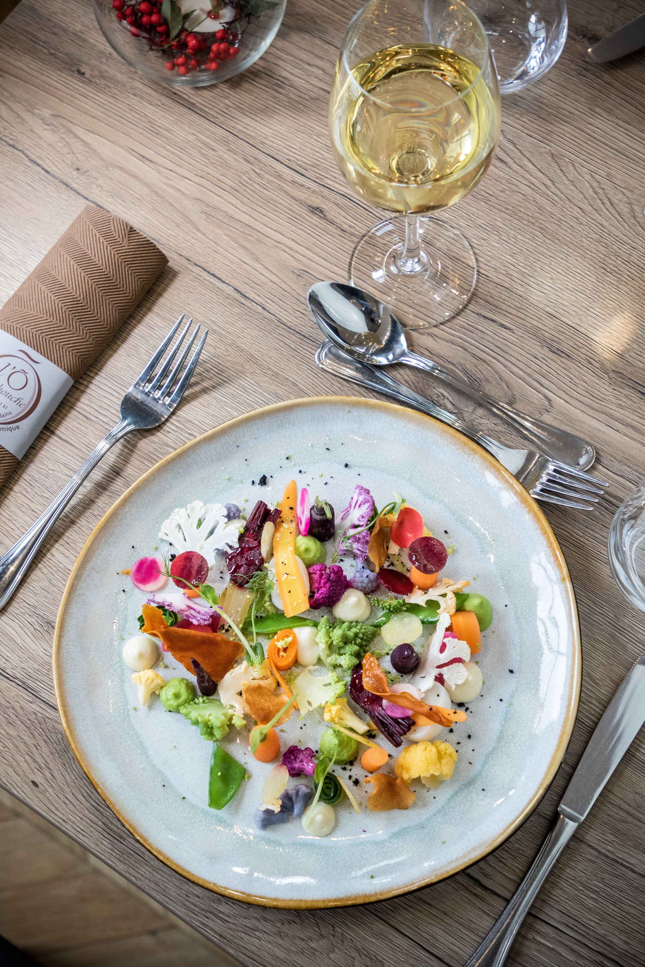 Une table à manger avec une assiette de salade colorée, un verre de vin blanc et des couverts.