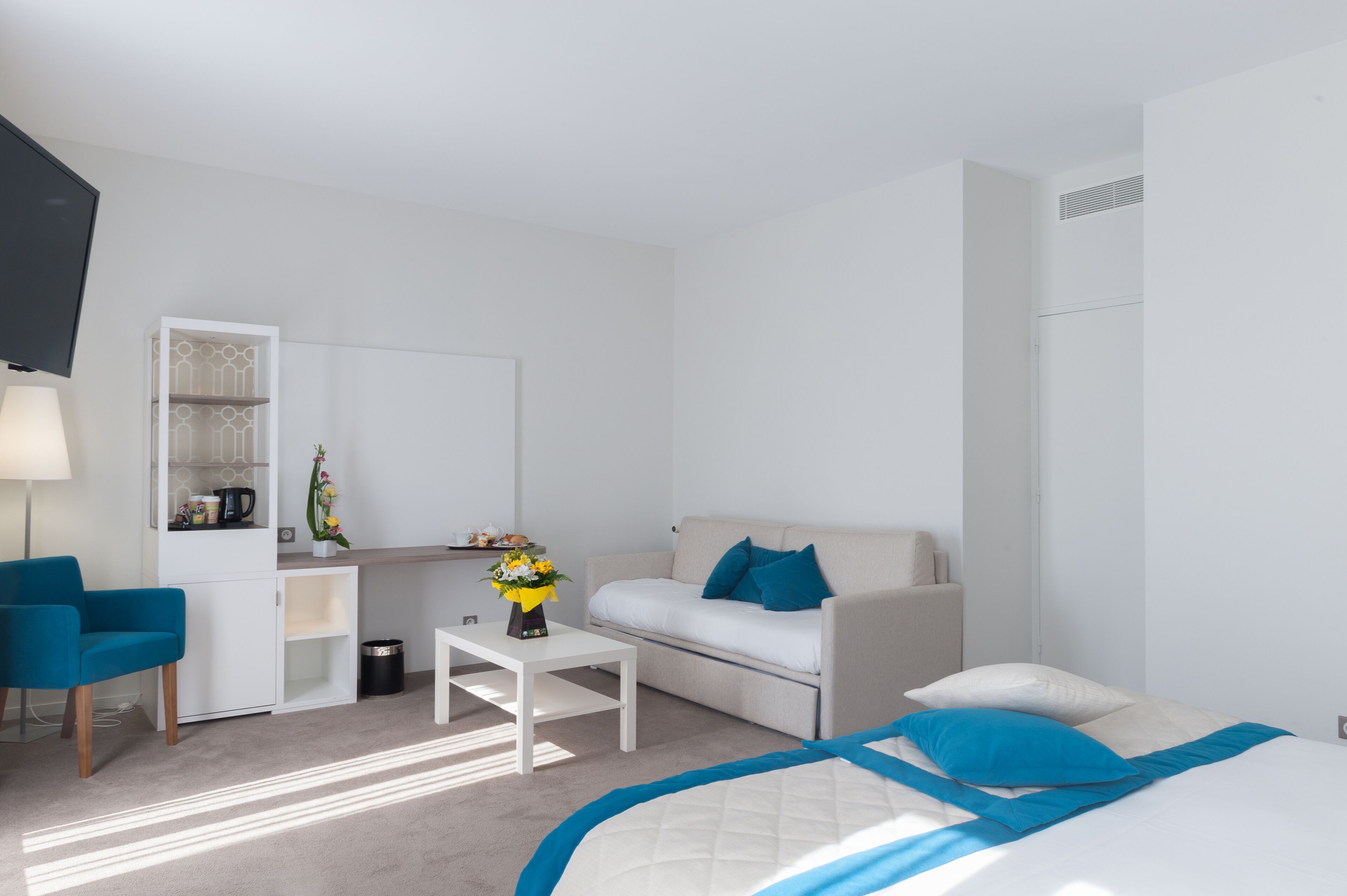 Una habitación de hotel moderna con cama, sofá, escritorio, silla, televisor y una kitchenette pequeña.