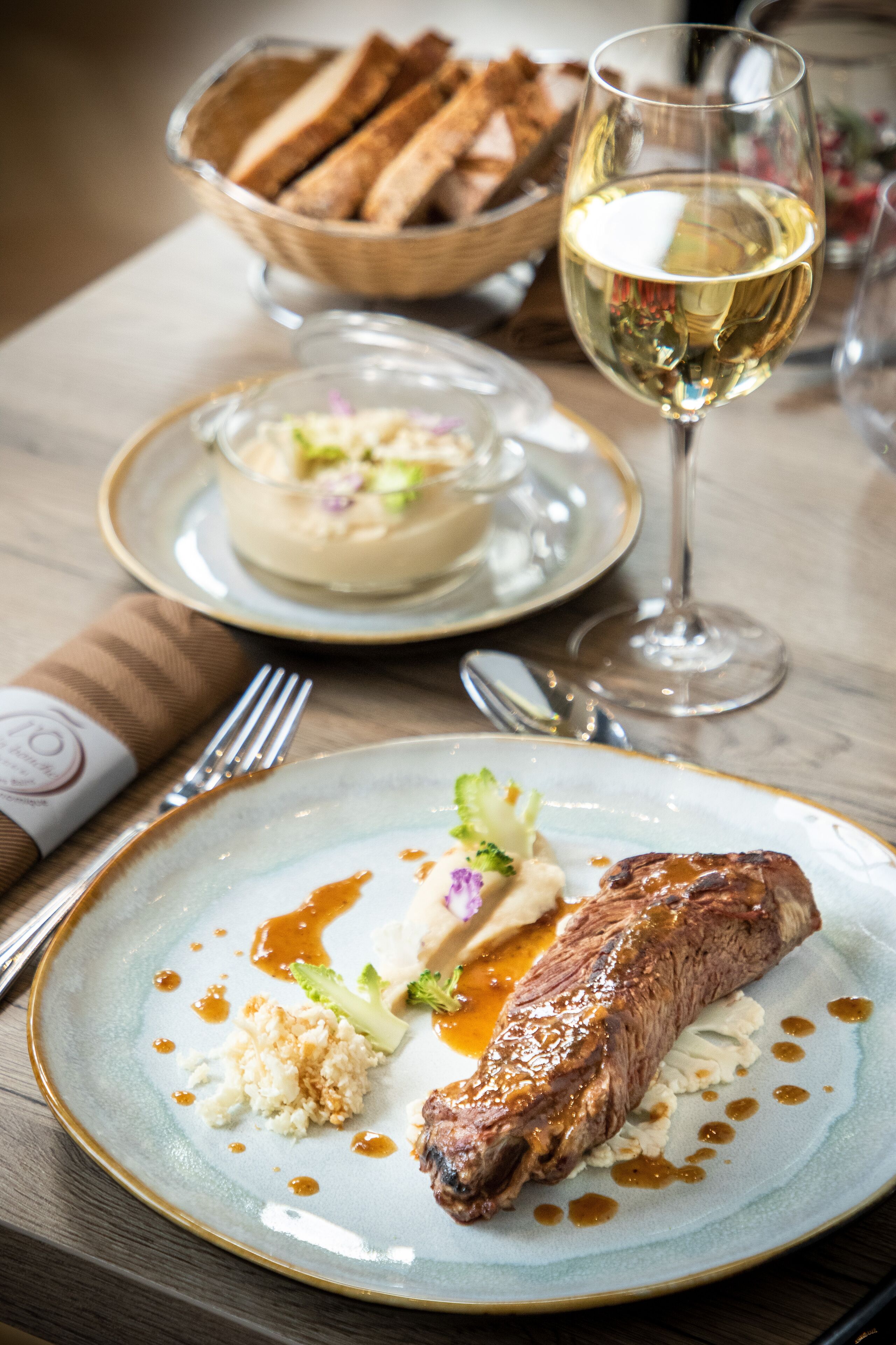 A dining table with a plate of steak, a glass of white wine, and a bowl of bread.