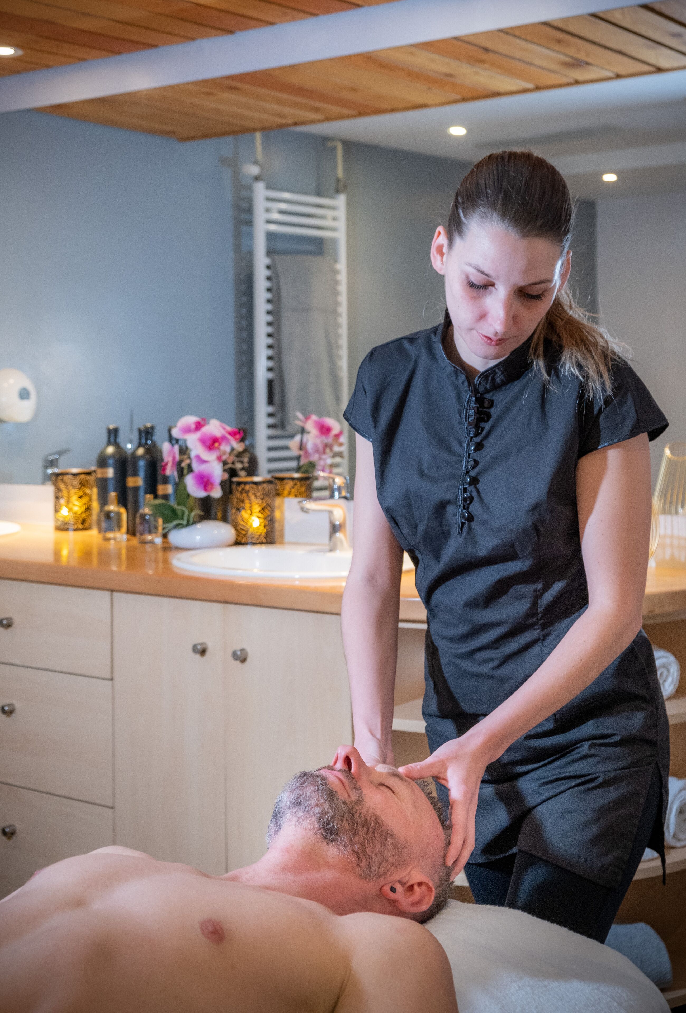 A man receiving a massage in a room with a wooden ceiling, a sink, and decorative bottles and flowers on the counter.