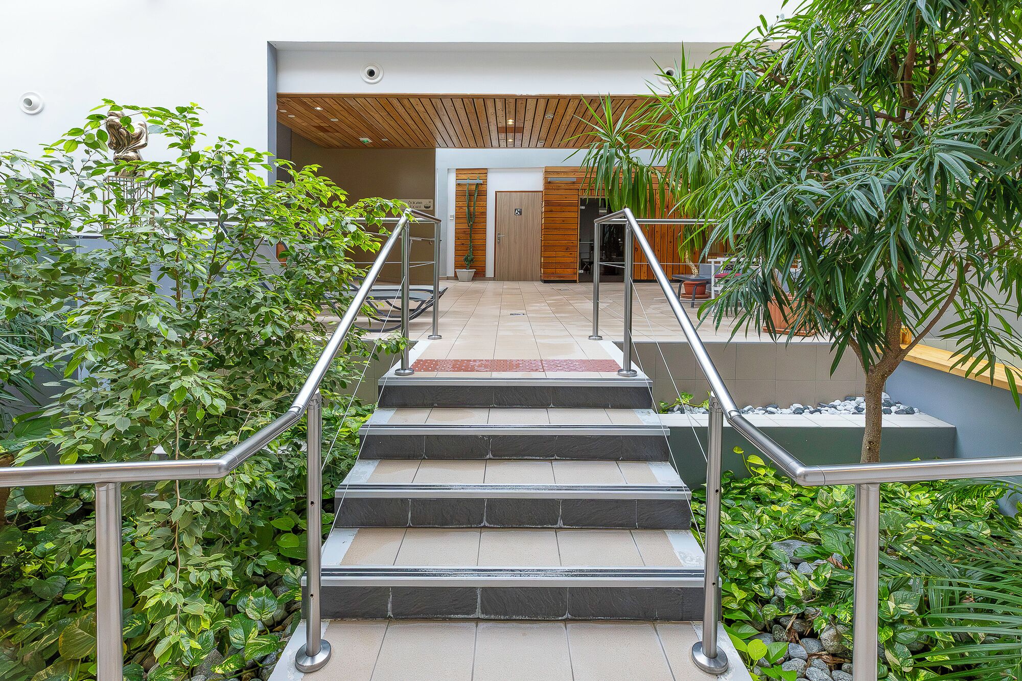 Una escalera conduce a la entrada de un edificio con barandales metálicos, rodeada de follaje y un techo de madera.