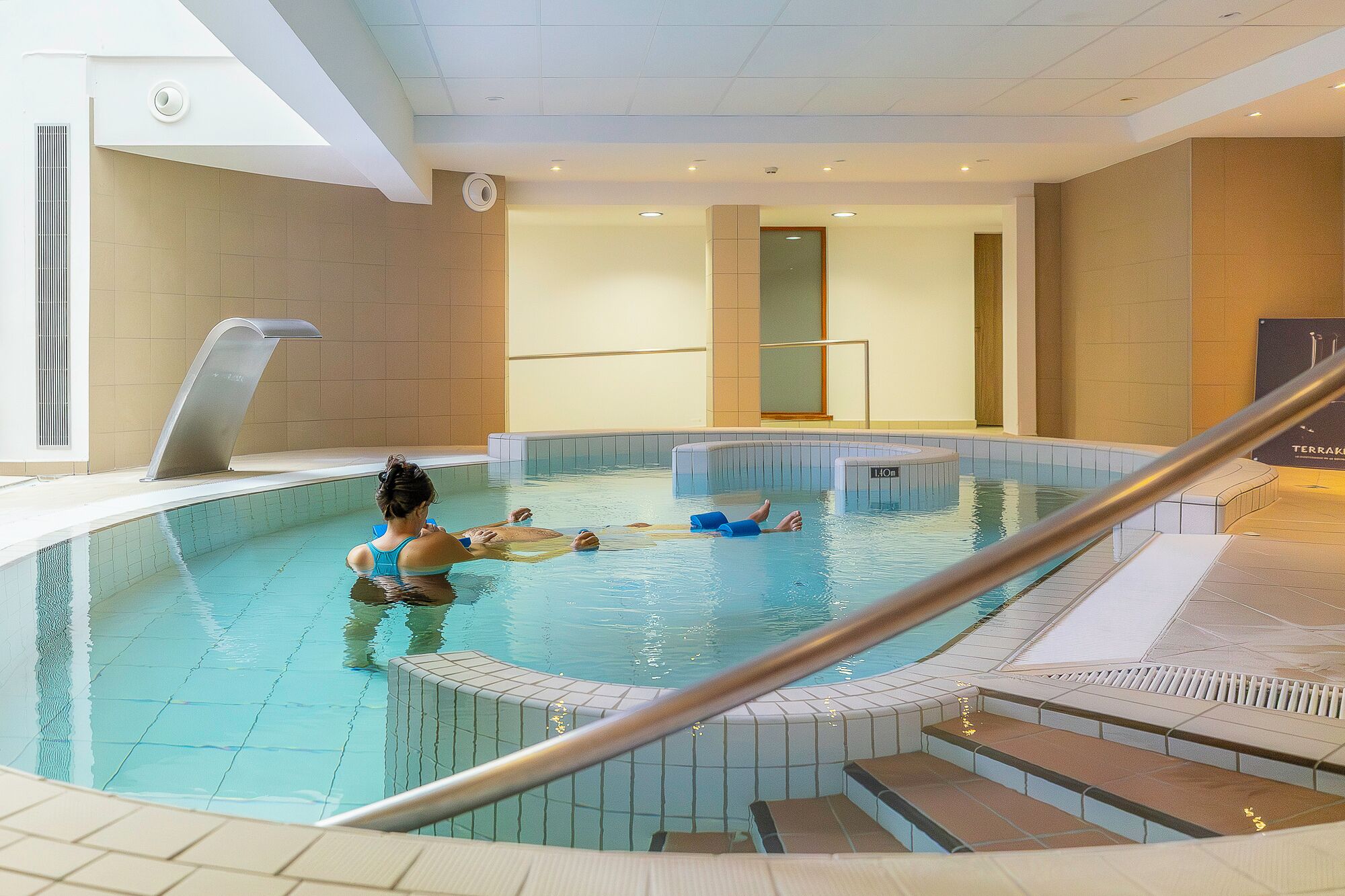 An indoor swimming pool with a person in the water, a pool ladder, and a tiled floor.