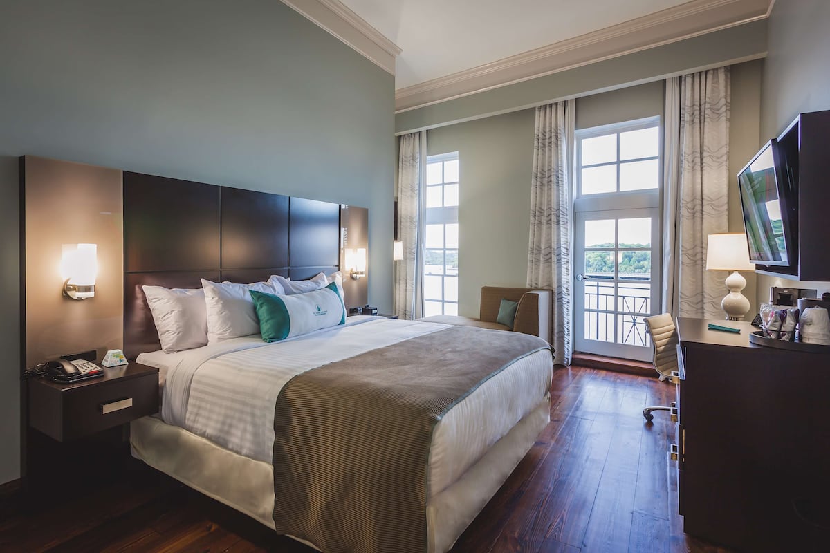 A hotel room with a large bed, bedside tables, a desk, a TV, and a balcony with curtains.